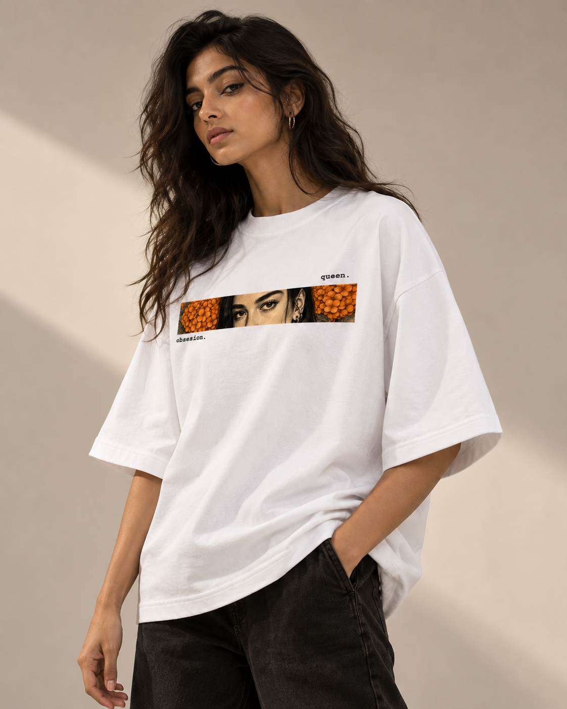 A woman wearing a white oversized graphic T-shirt with a rectangular chest print featuring a stylized face and orange circular patterns, paired with dark jeans against a warm beige studio background.