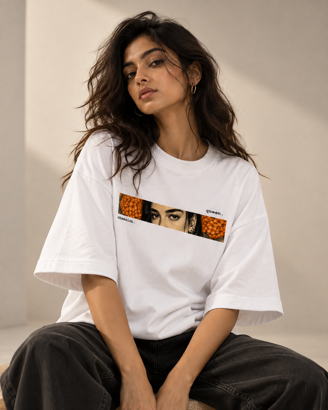 A woman wearing a white oversized graphic T-shirt with a rectangular chest print featuring orange patterned sections and a portrait, paired with dark wide-leg pants, seated against a warm beige wall.