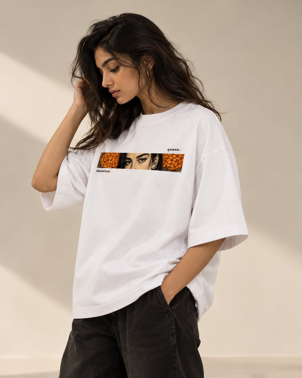 A woman wearing a white oversized T-shirt with a horizontal graphic print featuring a collage of faces and orange circular patterns, paired with dark gray jeans, set against a warm beige background with soft shadows.