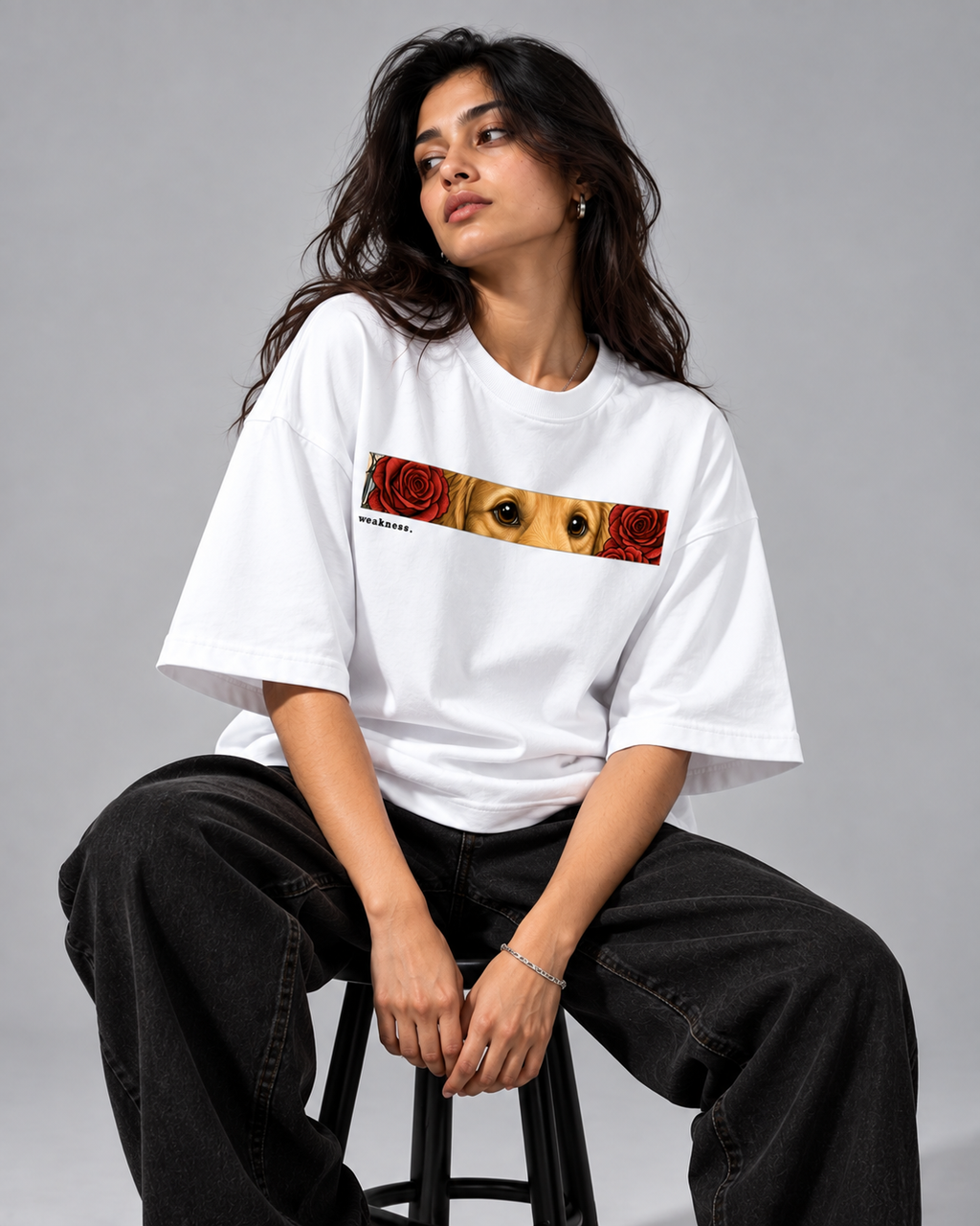 A woman sits on a stool wearing a white oversized graphic T-shirt with a horizontal print of a dog's face framed by red roses, paired with loose black jeans against a plain gray studio background.