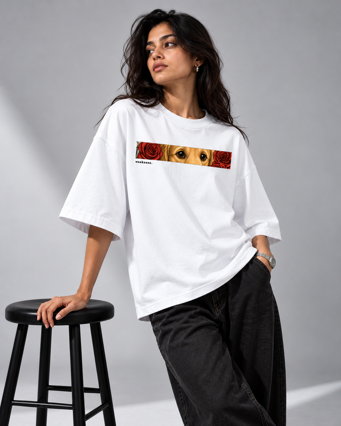 A woman wearing an oversized white graphic T-shirt with a horizontal rectangular print featuring a dog's face and red roses, paired with loose black pants, leaning on a black stool against a gray studio background.